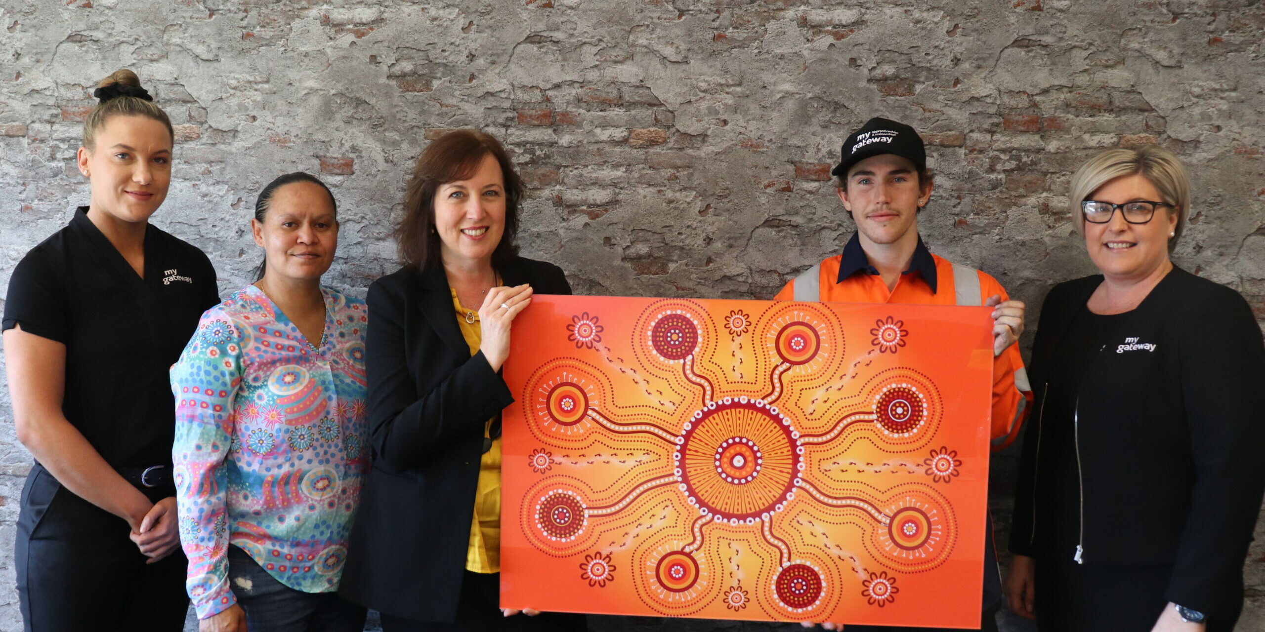 Five people stand in front of a mottled wall. A woman and a young man hold a painting between them. It is predominantly orange and yellow in colour with a circle at centre connected to other circles radiating from the first.