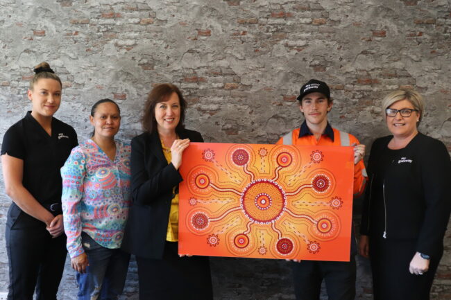Rhonda Sampson’s Indigenous designs captivate apprentice employers Five people stand in front of a mottled wall. A woman and a young man hold a painting between them. It is predominantly orange and yellow in colour with a circle at centre connected to other circles radiating from the first.
